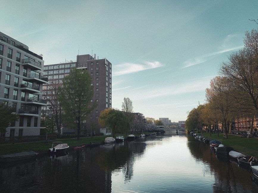 A picturesque canal scene in Amsterdam with modern buildings and moored boats under a clear blue sky.